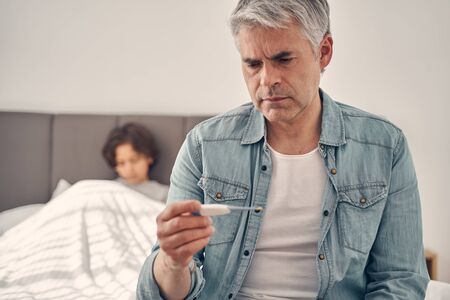 Grey haired man sitting in bedroom near his son while checking temperature on thermometer