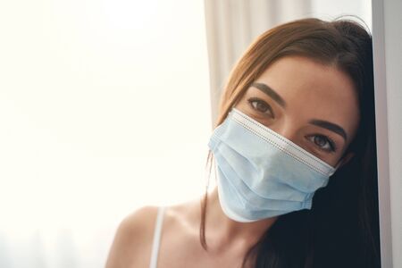 Close up portrait of young attractive dark-haired lady in protective mask standing near the wall...