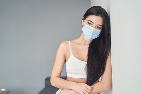 Attractive young female in white top and protective mask standing near the wall at home and looking awayの写真素材