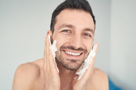 Happy cheerful male applying shaving soap on his cheeks and chin while looking at cameraの写真素材