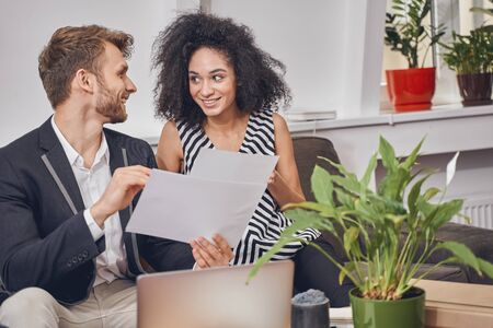 Cute young woman and her elegant male colleague with documents smiling at each otherの写真素材