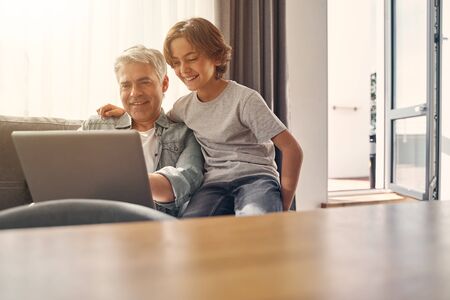 Happy child and grey haired father relaxing on couch while using laptop in living room at home