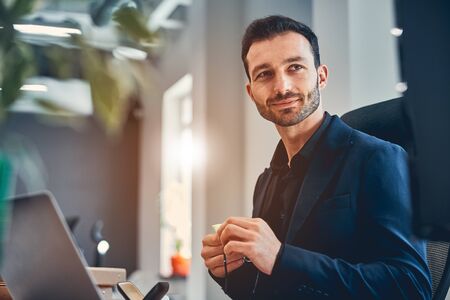 Happy bearded man in classic jacket wiping his glasses while working with computer