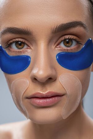 Close up portrait of calm young female wearing cosmetic masks under eyes and on nasolabial folds isolated on grey backgroundの写真素材