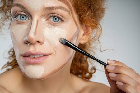 Close up portrait of smiling curly female using cosmetic and brush for improving her look isolated on grey backgroundの写真素材