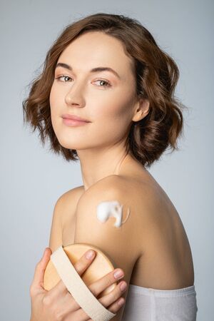 Portrait of cheerful curly female massaging body skin with soap and sponge isolated on grey backgroundの写真素材