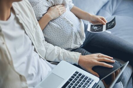 Close up of a pregnant woman holding a baby scan picture while her husband sitting with a laptop and putting a hand on her lapの写真素材