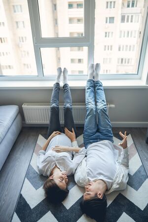 Lovely pregnant lady and her husband wearing similar clothes and lying on the carpet with their feet on a window sillの写真素材