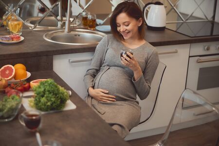 Cheerful pretty lady touching her belly and smiling to it while drinking tea in the kitchen