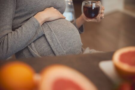 Cropped photo of a pregnant lady holding a glass mug of tea and putting one hand on the belly while sitting at the tableの写真素材