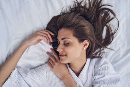 Close up of smiling pretty lady with closed eyes resting on white pillow in bedの写真素材