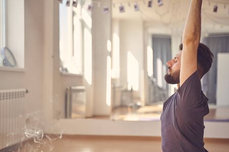 Serene young man closing eyes and raising hands while meditating during yoga practice