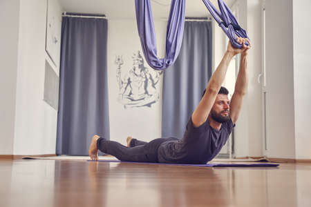 Bearded male lying on the floor and holding hammock swing while doing antigravity yoga exerciseの写真素材