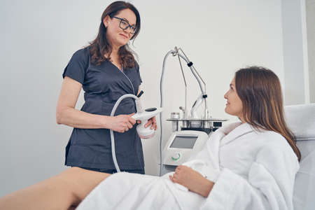 Gladsome professional cosmetician smiling to a client while holding a hand piece for ultrasound cavitationの写真素材