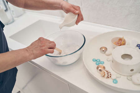 Hands of woman pouring water into the bowl with white powder clay and mixing it with a wooden stick