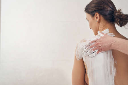 Caucasian woman sitting in beauty salon and cosmetologist spreading white clay on her back