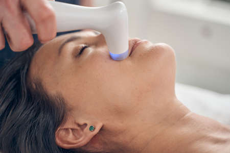 Young lady lying with her eyes closed and a professional cosmetologist applying low level laser to her skin on the cheekの写真素材