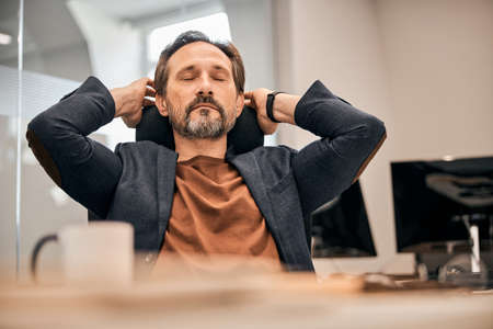 Close up portrait of gentleman in black jacket sitting on the chair with closed eye and putting hands upの写真素材