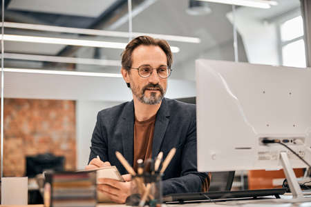Confident businessman in eyeglasses and brown shirt sitting at the table and looking at computer screen with serious facial expressionの写真素材