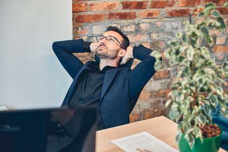 Successful project manager holding hand behind his head and enjoying moment while sitting at the modern workplaceの写真素材