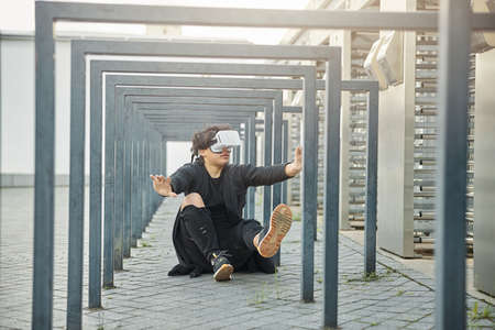 Stylish guy in black outfit sitting on asphalt and wearing virtual reality glasses