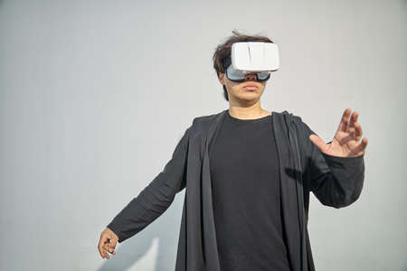 Stylish guy in black outfit standing against white wall and using VR headset