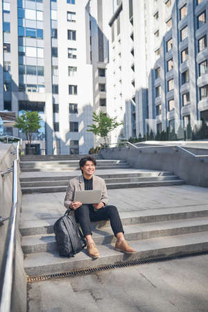 Handsome stylish guy with laptop looking away and smiling while sitting on steps outdoors