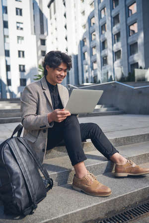 Cheerful guy in stylish jacket surfing the internet on notebook and smiling while resting on street...