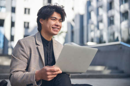 Cheerful guy in stylish jacket looking away and smiling while holding modern laptopの写真素材