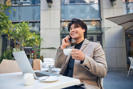 Handsome guy with cup of hot drink talking on mobile phone and smiling while sitting at the table with laptopの写真素材