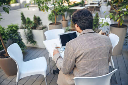 Back view of stylish guy sitting at the table with cup of coffee and modern laptop