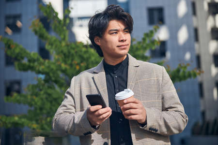 Handsome stylish guy holding takeaway cup of coffee and smartphone while standing on the street