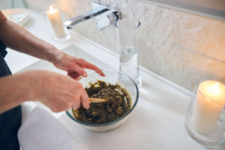 Hands of beautician mixing substance in a glass bowl while standing by the sink with candles burning nearの写真素材