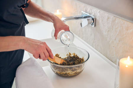 Unrecognizable spa salon worker standing next to the sink and mixing clay with water for the...