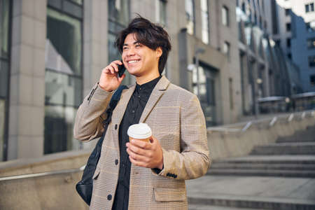 Handsome guy in jacket holding takeaway cup of coffee and smiling while having phone conversation outdoorsの写真素材