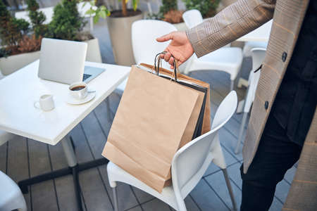 Close up of male hand holding paper bags with purchases while gentleman standing near table with laptop and cup of coffeeの写真素材