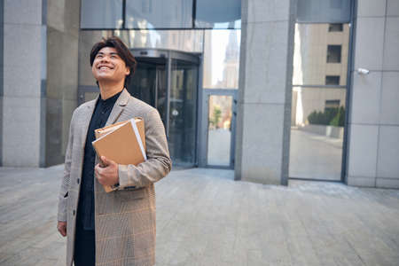 Handsome stylish guy looking up and smiling while holding folder and spiral sketchbookの写真素材