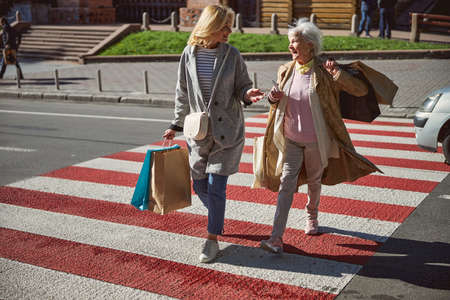 Joyful female friends walking in street and holding bright paper shopping bags while talking and enjoying conversationの写真素材