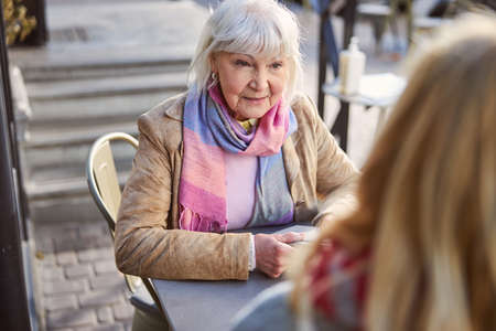 Beautiful smiling old lady wearing pink scarf sitting in the restaurant terrace with cup of coffee while talking with another femaleの写真素材