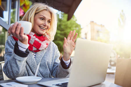 Close up portrait of happy nice lady holding mug of tea in hand while looking the screen grey laptop