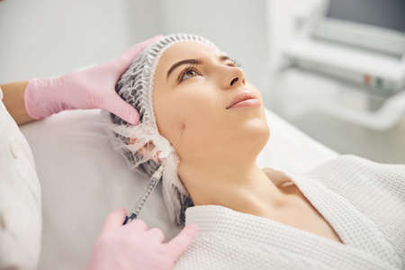 Calm female patient getting face lifting injection while lying on couch and looking upwardsの写真素材
