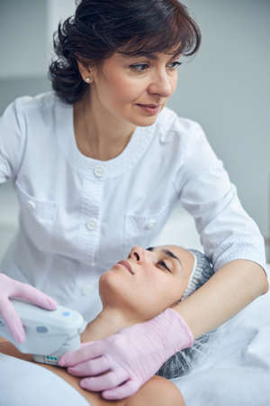 Mature beauty expert using modern apparatus while looking away and performing non surgical...