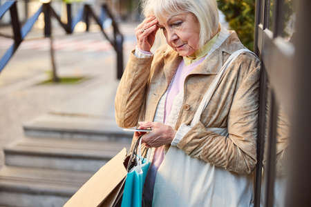 Elderly woman thinking about something while holding mobile phone and shopping bags in her handsの写真素材