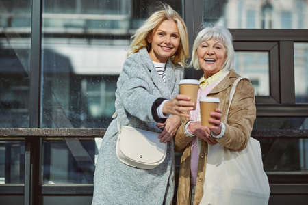 Laughing cheerful ladies standing at the modern street of big city while drinking hot drinksの写真素材