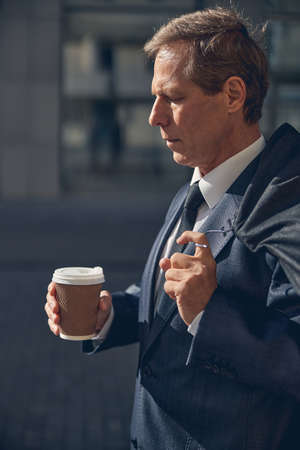 Elegant gentleman looking at cup of coffee with serious expression while holding suit garment bag