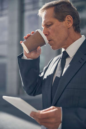 Serious gentleman in suit holding electronic pad PC and drinking takeaway hot beverage on the streetの写真素材
