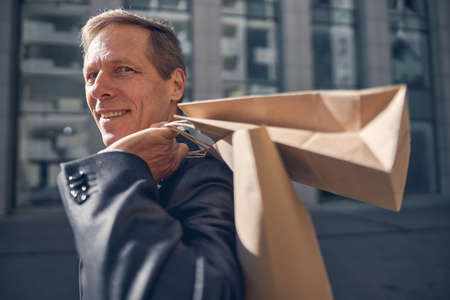 Close up of handsome gentleman looking at camera and smiling while carrying shopping bags on his...