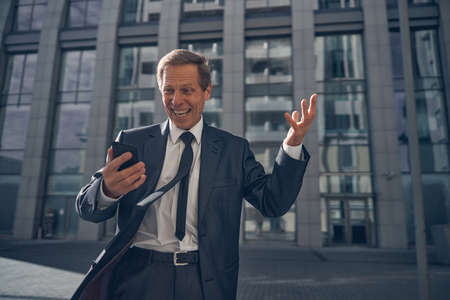 Joyful happy businessman reading message with good news on smartphone while standing on the street