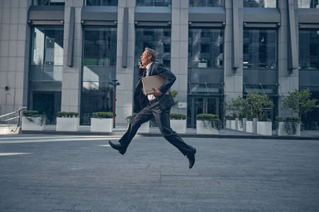 Joyful businessman in elegant suit talking on cellphone and smiling while holding documents and running outdoorsの写真素材