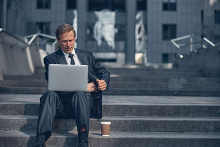 Imposing serious man in elegant suit sitting on steps and working on notebook outdoors